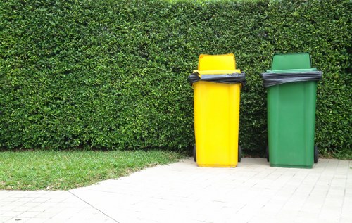 Two plastic wheelie bins, one yellow and one green, are positioned on a paved pathway in front of a dense, neatly trimmed green hedge. The yellow bin is slightly taller and leans slightly to the left, with a black lid resting on top. The green bin, with a closed matching lid, stands directly beside it. Both bins are situated on the paved surface, which appears clean and well-maintained, adjacent to a strip of well-kept grass. The background features the thick hedge, forming a natural backdrop that emphasizes the presence of waste bins typically used for rubbish collection services in the area. The scene is evenly lit, suggesting daylight conditions, and presents a clear, straightforward view relevant for a rubbish removal company's website, such as Swiss Cottage House Clearance, serving the NW3 postcode area. The setting implies a residential environment suitable for domestic rubbish disposal and clearance activities.