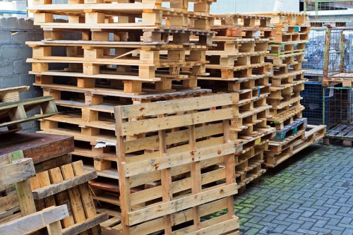 A large stack of wooden pallets arranged in multiple columns on a paved outdoor surface, with some pallets leaning against the main pile. The pallets are made of light-colored, unfinished timber with visible wood grain and rough edges, and they appear weathered with some staining and minor damage. The background includes a section of a brick wall, parts of a metal fence, and an urban environment. The overall scene suggests a storage or waste collection area for timber pallets, consistent with rubbish removal services. The image provides a clear view of the neatly stacked pallets intended for disposal or recycling, relevant to Swiss Cottage House Clearance’s local rubbish clearance activities in the area.