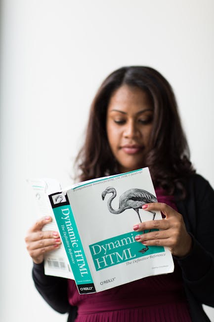 A woman with dark, shoulder-length hair and medium skin tone is standing indoors against a plain white background. She is wearing a dark blazer over a maroon top. The woman is holding a book titled 'D